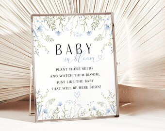Op de afbeelding: Een witte kaart met blauwe bloemenmotieven en de tekst "BABY in bloom" met een hartje. De kaart bevat ook de tekst "PLANT THESE SEEDS AND WATCH THEM BLOOM, JUST LIKE THE BABY THAT WILL BE HERE SOON!" met een hartje.