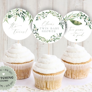 以下が含まれることがあります： 緑の葉と白い花がついたカップケーキの飾り3つ。飾りには「It's Twins!」「Olivia's Twin Baby Shower」「Two Peas in a Pod」と書かれています。