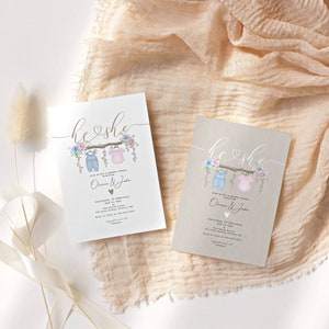 May include: Two gender reveal party invitations with a rustic brown background. The invitations feature a watercolor illustration of a clothesline with a blue baby outfit and a pink baby outfit hanging on it. The text "he & she" is written in cursive above the clothesline. The invitations also include the text "Join us for a gender reveal party" and the names "Olivia & Jake".