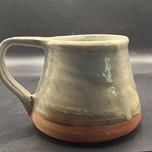 Può includere: Tazza in ceramica fatta a mano con corpo arrotondato e grande manico. La tazza presenta un design bicolore, con una parte superiore verde tenue e una base marrone rossiccio. La superficie della tazza ha un aspetto leggermente strutturato.