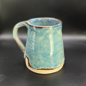 May include: Handmade ceramic mug with a light blue glaze. The mug has a rounded body, a sturdy handle, and a contrasting brown rim. The base is a natural, unglazed colour. A unique piece for enjoying coffee or tea.