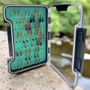 Może przedstawiać: Otwarta skrzynka na muchy wędkarskie z przezroczystą pokrywą, odsłaniająca zielone wnętrze wypełnione różnymi kolorowymi muchami wędkarskimi. Muchy są starannie ułożone, prezentując gamę kolorów i stylów, gotowe do użycia. Pudełko znajduje się na kamiennej powierzchni.