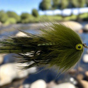 Könnte beinhalten: Nahaufnahme einer handgefertigten olivgrünen und schwarzen Fliegenfisch-Köder. Der Köder hat ein gelbes Auge und soll einen kleinen Fisch imitieren. Die Fliege besteht aus Federn und anderen Materialien und soll Fische anlocken.