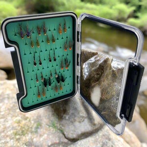 Può includere: Una scatola da pesca a mosca nera con un coperchio trasparente, aperta per rivelare un inserto in schiuma verde con file di mosche colorate. La scatola è appoggiata su uno scoglio vicino a un torrente.