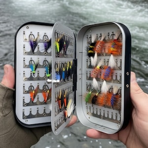May include: An open, rectangular fly fishing tackle box with a black frame and silver interior. The box is filled with various colorful fishing flies, including orange, yellow, blue, and black. The box is held open, revealing the contents.