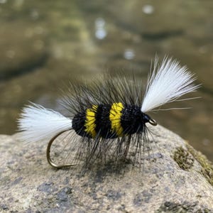Puede incluir: Una mosca de pesca artesanal con cuerpo negro, rayas amarillas y detalles de plumas blancas. La imitación de insecto tiene un anzuelo curvo y está sobre una piedra gris cerca del agua.