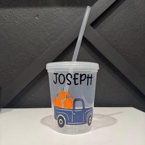 May include: Clear plastic tumbler with a lid and straw. The tumbler features the name "JOSEPH" in black lettering. A blue truck graphic carries orange pumpkins. A fall-themed cup for beverages.