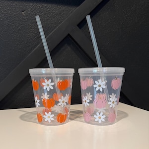 May include: Two clear plastic tumblers with lids and straws. One tumbler features orange pumpkins and white daisy flowers, the other has pink pumpkins and white daisies. The tumblers are on a white surface.