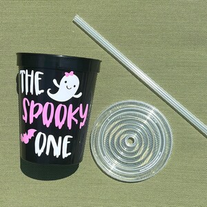 May include: Black reusable cup with a white ghost and the text "The Spooky One" in pink. The cup comes with a clear lid and a clear straw.