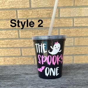 May include: Black reusable cup with a white ghost and bat graphic and the text "The Spooky One" in pink lettering. The cup has a clear plastic straw.