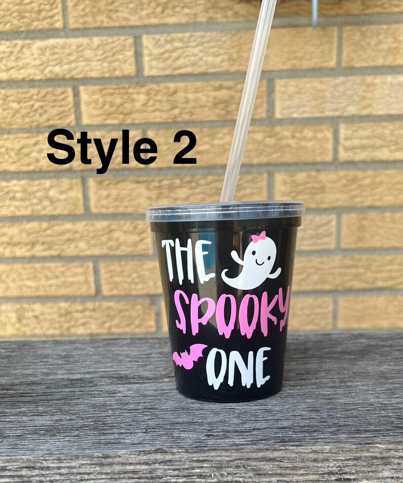 May include: Black reusable cup with a white ghost and bat graphic and the text "The Spooky One" in pink lettering. The cup has a clear plastic straw.