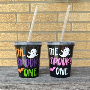 May include: Two black reusable cups with clear straws. The cups have a white ghost with a pink bow and the text "The Spooky One" in pink and orange.