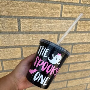 May include: Black reusable cup with a white ghost and pink bow, and the text "The Spooky One" in pink and white lettering. A clear plastic straw is in the cup.