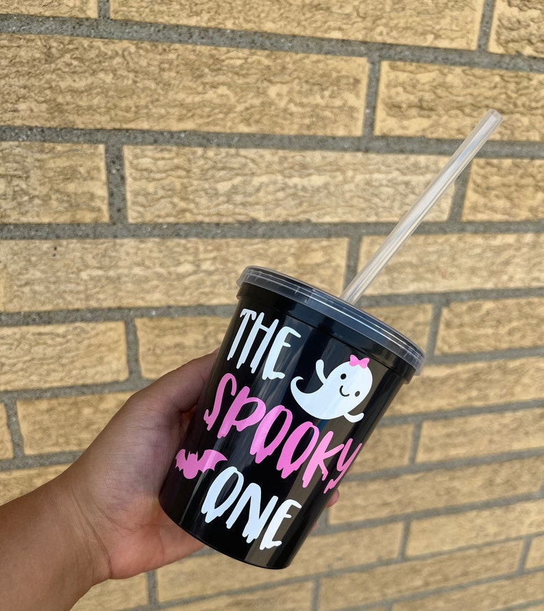 May include: Black reusable cup with a white ghost and pink bow, and the text "The Spooky One" in pink and white lettering. A clear plastic straw is in the cup.