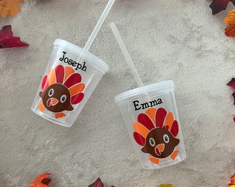 May include: Two clear plastic cups with straws, featuring a Thanksgiving turkey design and personalized names "Joseph" and "Emma". The cups are on a white background with fall leaves.