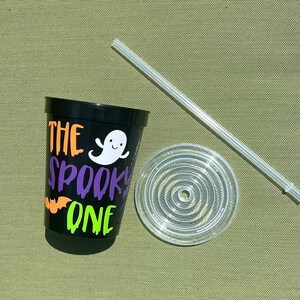 May include: Black reusable cup with the text "The Spooky One" in orange, green, and purple lettering. A white ghost with a smiling face is next to the text. A clear plastic lid and straw are next to the cup.