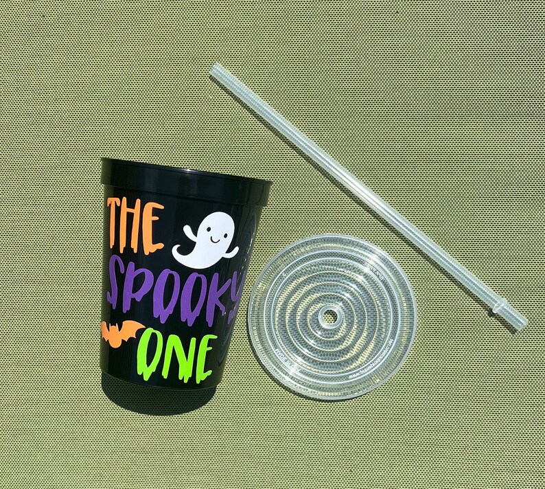May include: Black reusable cup with the text "The Spooky One" in orange, green, and purple lettering. A white ghost with a smiling face is next to the text. A clear plastic lid and straw are next to the cup.