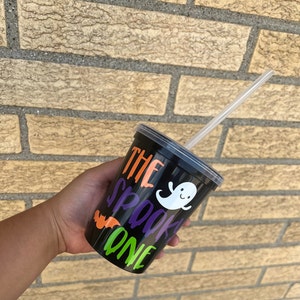 May include: Black reusable cup with a clear lid and a straw. The cup has a ghost and bat design with the text "The Spooky One" in orange, purple, and green.