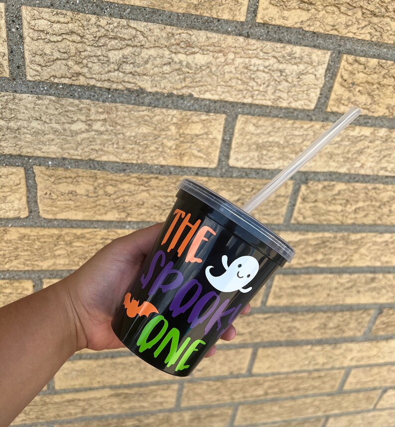 May include: Black reusable cup with a clear lid and a straw. The cup has a ghost and bat design with the text "The Spooky One" in orange, purple, and green.