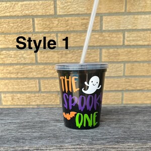 May include: Black plastic cup with a clear plastic straw. The cup has a design that says "The Spooky One" with a ghost, bat, and dripping green and orange paint.