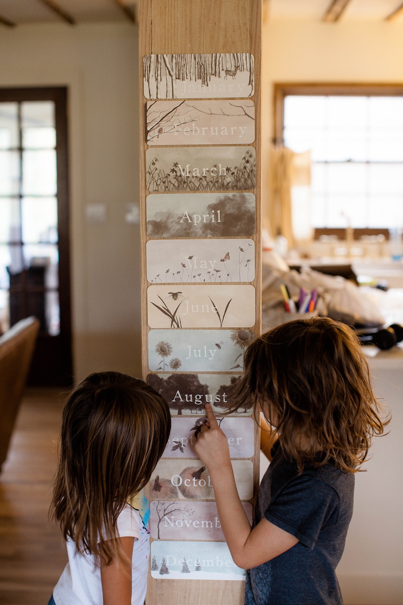 May include: Two children are looking at a wooden calendar with images of nature for each month. The calendar has the months of February, March, April, May, June, July, August, September, October, November, and December.