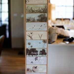May include: A wooden wall with a set of twelve decorative cards featuring illustrations of nature scenes and the months of the year. The cards are labeled with the months: February, March, April, May, June, July, August, September, October, November, and December.