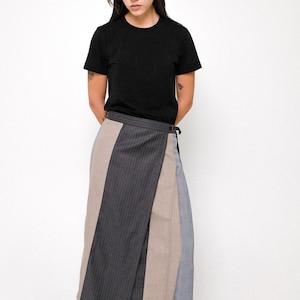 May include: A black short-sleeved t-shirt paired with a midi-length wrap skirt featuring vertical panels of charcoal pinstripe, solid beige, and light blue fabric. The skirt includes a side buckle closure and is worn with black leather boots.