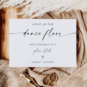 May include: A white sign with black script that reads "LIGHT UP THE dance floor. HELP YOURSELF TO A glow stick." A gold wax stamp and gold wax seals are at the bottom. The sign is on a wooden tray with beige fabric.