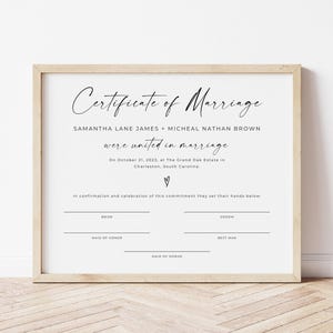 May include: A framed marriage certificate with stylish calligraphy. The text reads "Certificate of Marriage" above the couple's names and the wedding date and location. The frame is a light wood colour.