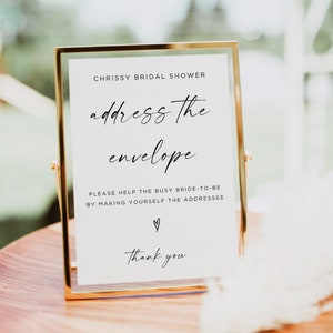 May include: A gold-framed sign with white text that reads "Chrissy Bridal Shower address the envelope Please help the busy bride-to-be by making yourself the addressee ♡ Thank you".