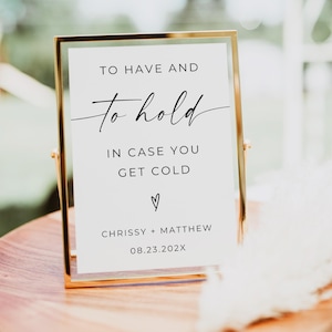 May include: A gold-framed sign with a white background and black text that reads "To have and to hold, in case you get cold. ♡ Chrissy + Matthew 08.23.202X".