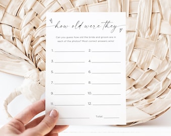 May include: A white card with the text "how old were they" and lines for answers. The card is for a wedding game, asking guests to guess the bride and groom's ages in photos. The card is held in a hand.