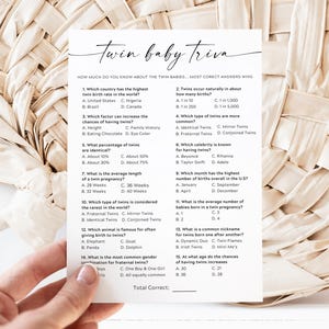 May include: A white card with the text "Twin baby trivia" in elegant script. The card features multiple-choice questions about twins, held by a hand against a woven background. A fun game for a baby shower.
