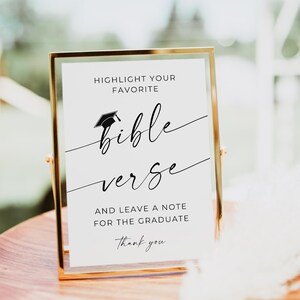 May include: A vertical rectangular sign in a thin gold metal frame. The white sign features black text that reads: HIGHLIGHT YOUR FAVORITE bible verse AND LEAVE A NOTE FOR THE GRADUATE Thank you. A small black graduation cap icon sits above the word bible.