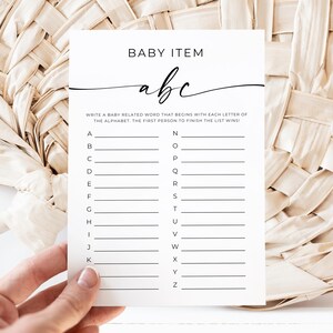 May include: A white baby shower game card with the text "BABY ITEM" and "abc". The card has lines for writing baby-related words, one for each letter of the alphabet. The card is held by a hand.