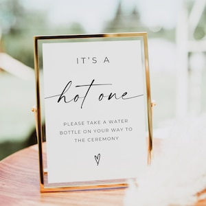 May include: A gold framed sign with the text "IT'S A hot one" in black script. Below, it reads "PLEASE TAKE A WATER BOTTLE ON YOUR WAY TO THE CEREMONY" with a heart symbol. The sign is on a wooden surface.