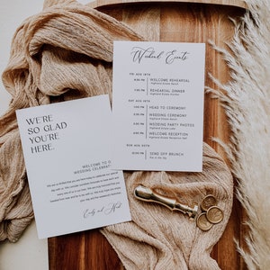Op de afbeelding: Een witte kaart met zwarte tekst die "We're so glad you're here." en "Welcome to our wedding celebration!" zegt. De kaart ligt op een houten oppervlak met een beige stof en gouden waszegels.