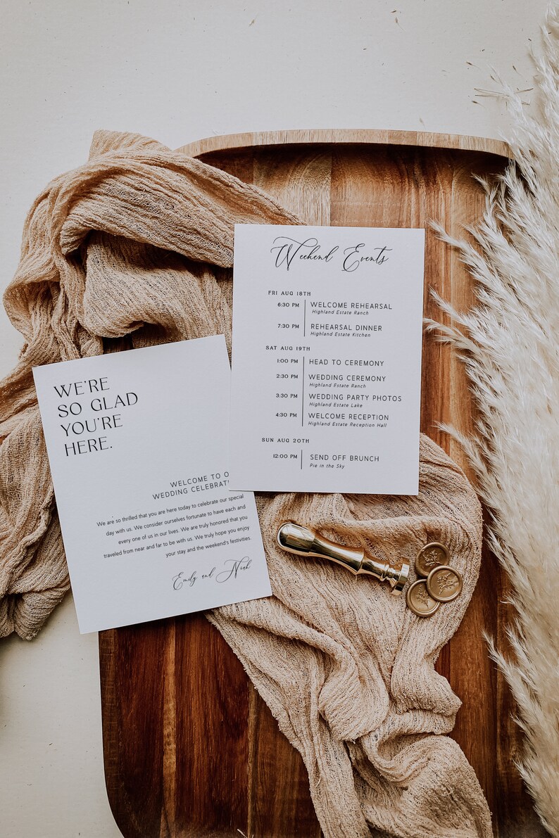 Op de afbeelding: Een witte kaart met zwarte tekst die "We're so glad you're here." en "Welcome to our wedding celebration!" zegt. De kaart ligt op een houten oppervlak met een beige stof en gouden waszegels.