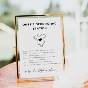 Op de afbeelding: Een ingelijst bord met gouden rand toont instructies voor een onesie-decoratiestation. Het bord bevat de tekst "ONESIE DECORATING STATION" en stappen voor het aanbrengen van vinylontwerpen op onesies. Het bord is voor een "baby christopher shower."