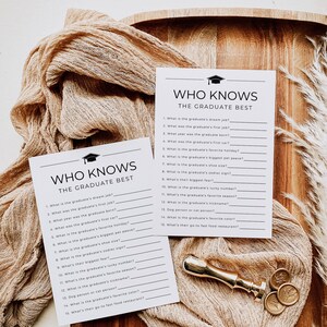Op de afbeelding: Twee spelkaarten voor een afstudeerfeest met de tekst "Who Knows The Graduate Best" worden weergegeven op een houten oppervlak. Een gouden wasstempel en decoratieve stof zijn ook aanwezig. De kaarten bevatten invulvragen.
