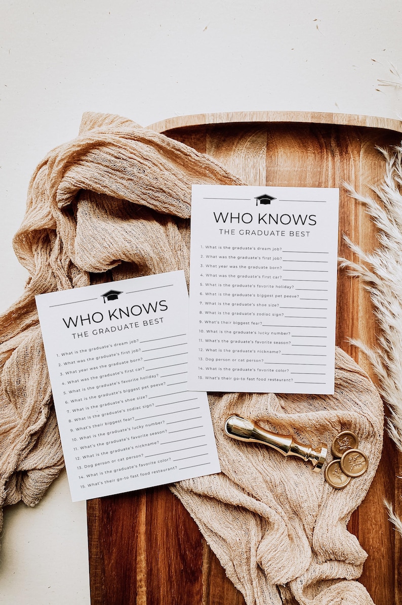 Op de afbeelding: Twee spelkaarten voor een afstudeerfeest met de tekst "Who Knows The Graduate Best" worden weergegeven op een houten oppervlak. Een gouden wasstempel en decoratieve stof zijn ook aanwezig. De kaarten bevatten invulvragen.