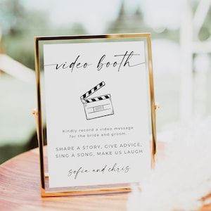 May include: A gold-framed sign with the text "video booth" in black cursive script. A black and white illustration of a clapperboard is below the text. The sign also includes the text "Kindly record a video message for the bride and groom. SHARE A STORY, GIVE ADVICE, SING A SONG, MAKE US LAUGH Sofia and chris".