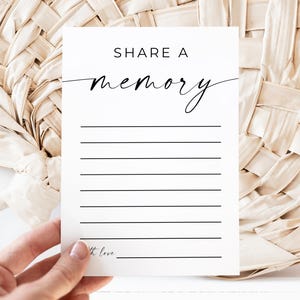 May include: A white card with the text "SHARE A memory" in black script. The card has lined spaces for writing and is held by a hand. The background features a woven, beige material.