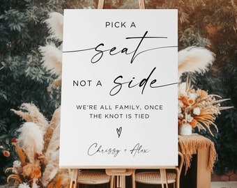 May include: White sign with black text that reads "Pick a seat, not a side. We're all family, once the knot is tied. ♡ Chrissy + Alex". The sign is on an easel in front of a floral arrangement.