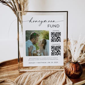 Peut inclure: Une pancarte blanche avec du texte noir qui dit "honeymoon fund" avec une photo d'un couple et des codes QR pour Venmo et CashApp. La pancarte est dans un cadre dor&eacute; et pos&eacute;e sur une table avec un vase brun et des fleurs s&eacute;ch&eacute;es.