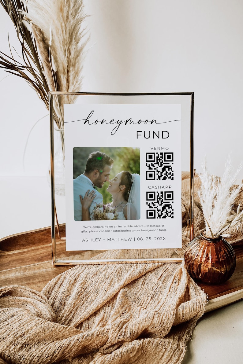 Peut inclure: Une pancarte blanche avec du texte noir qui dit "honeymoon fund" avec une photo d'un couple et des codes QR pour Venmo et CashApp. La pancarte est dans un cadre dor&eacute; et pos&eacute;e sur une table avec un vase brun et des fleurs s&eacute;ch&eacute;es.