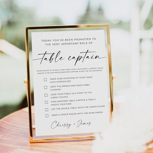 Könnte beinhalten: Ein rechteckiges Schild mit Goldrahmen und weißem Papier, das schwarzen Text enthält: TODAY YOU'VE BEEN PROMOTED TO THE VERY IMPORTANT ROLE OF Table captain. Es listet sechs Aufgaben für Gäste auf und ist unten mit Chrissy + James signiert.