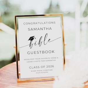 May include: A gold framed sign with the text "Congratulations Samantha Bible Guestbook". The sign includes a graduation cap graphic and the text "Class of 2026". The sign is on a wooden tray.
