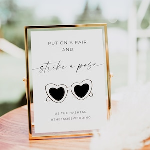 May include: A gold-framed sign with the text "PUT ON A PAIR AND strike a pose" and a graphic of heart-shaped sunglasses. The sign also includes the hashtag #THEJAMESWEDDING.