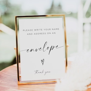 May include: A rectangular sign in a thin gold metal frame with white background and black text that reads: PLEASE WRITE YOUR NAME AND ADDRESS ON AN envelope, followed by a small hand-drawn heart and the words Thank you in script font.